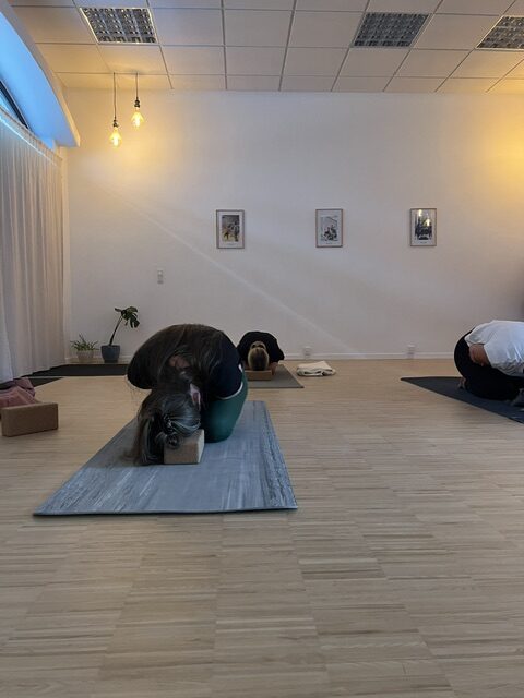 yin yoga esbjerg. 3 kvinder i forskellig alder laver 'kram barnet'. childs pose, med armene på tværs over maven. der er ekstra udstyr, en hvid væg og trægulv. Det emmer af fred og ro.